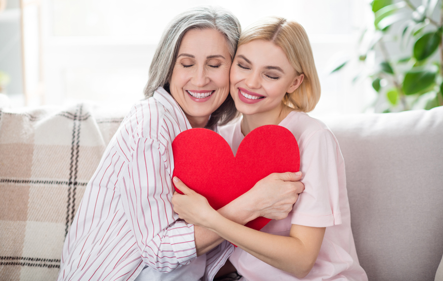 Muttertag: Ältere Mutter mit ihrer Tochter sitzen eng nebeneinander auf einer Coach und umarmen gemeinsam ein rotes Herz
